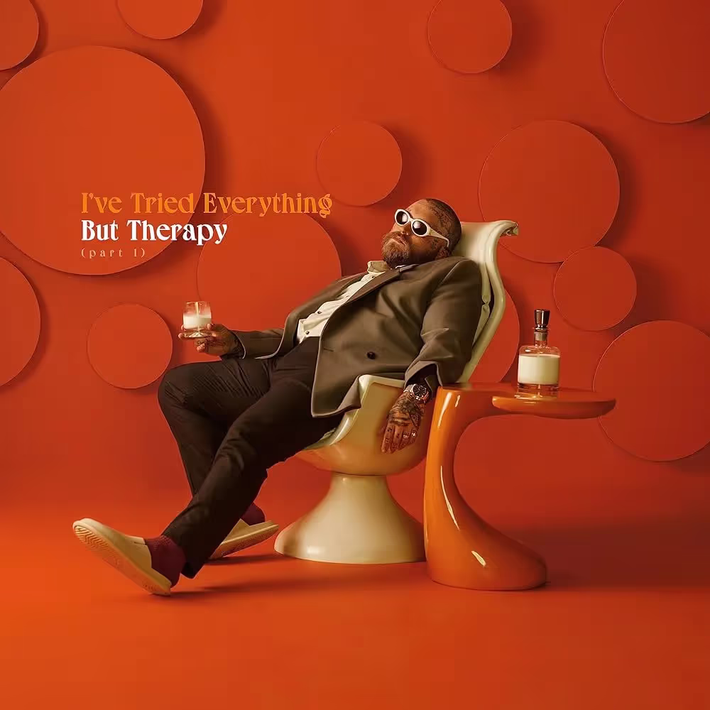 Teddy Swims lounging in a chair, holding what appears a glass of milk on a vibrant orange background for album cover of "Ive tried everything, but therapy"
