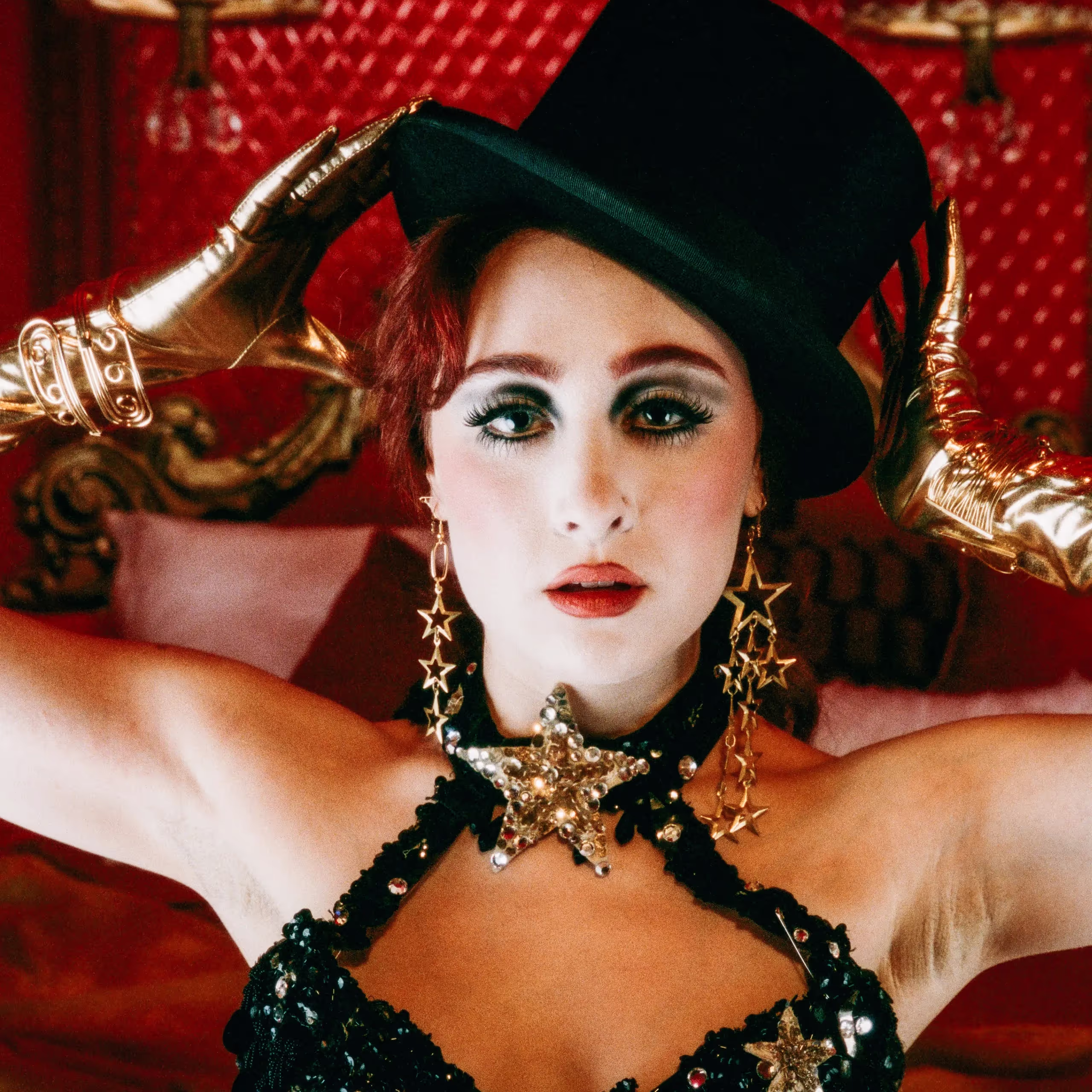 Chappell Roan poses for 'Red Wine Supernova' wearing a sparkling bra, green cylinder hat, and gold star earrings against a bright red background.