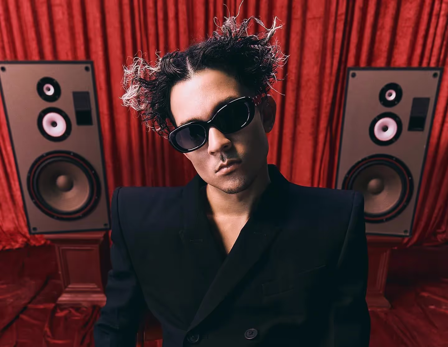 Tainy wearing black suit and sunglasses, on a red backdrop