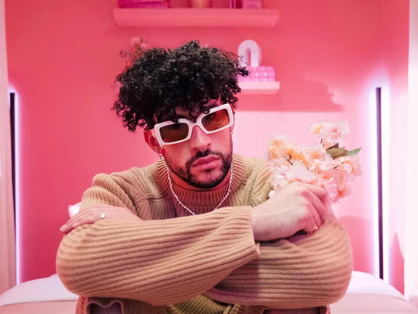 Bad bunny rocks natural curls, wearing white-frame sunglasses and cozy sweater, on a pink background.