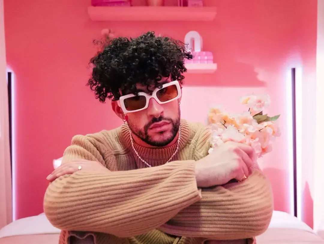 Bad bunny rocks natural curls, wearing white-frame sunglasses and cozy sweater, on a pink background.