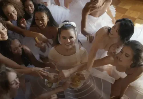 Selena Gomez wearing white lacy dress, surrounded by dancers in 'Love On' music video, released February 22, 2024