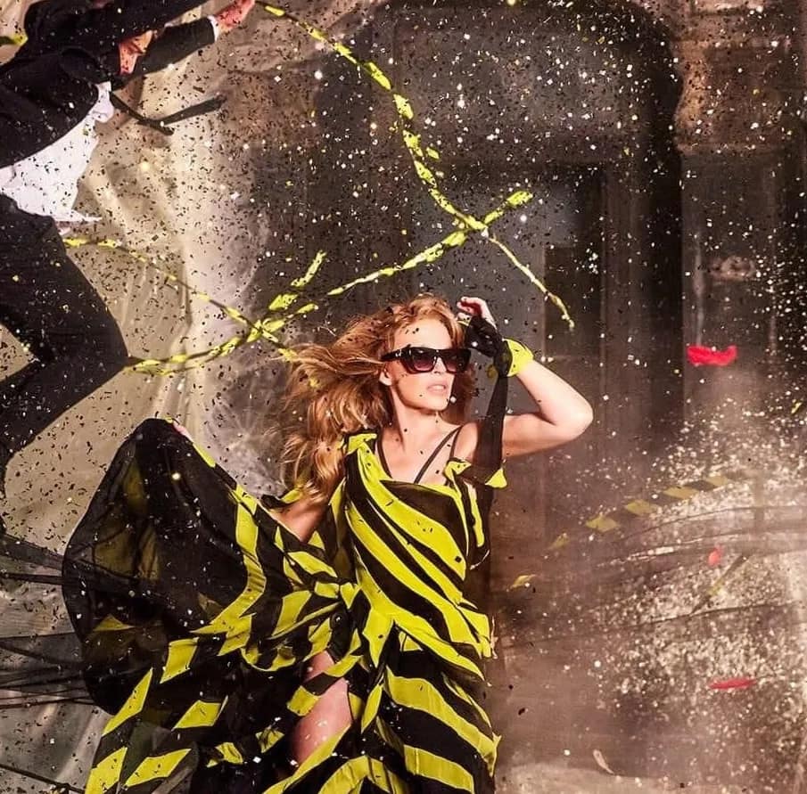 Kylie minogue in yellow and black striped dress, wearing glasses on set of camera, lights, action music video