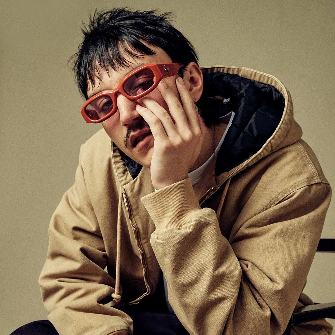 Artemas with edgy black haircut, orange glasses, brown jacket on olive green background.