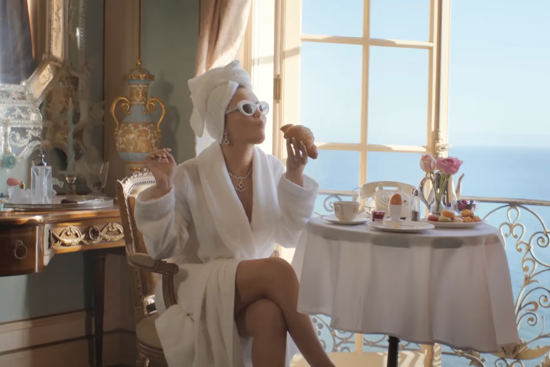 Selena Gomez dressed in a robe and jewelry, eating croissant in a luxury coastal hotel